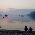 Smoky sky with boats on the water below and two children crouching on the beach.